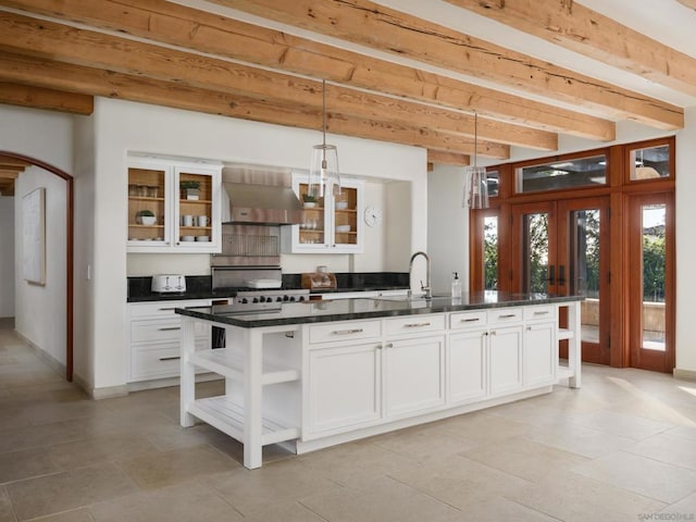 kitchen with wall chimney range hood, a kitchen island with sink, white cabinetry, beam ceiling, and hanging light fixtures