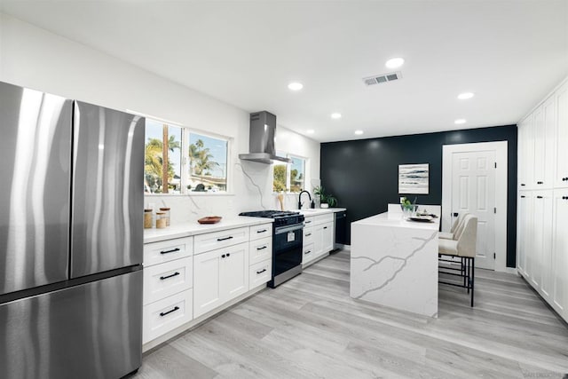 kitchen with wall chimney exhaust hood, light stone counters, a kitchen island, stainless steel appliances, and white cabinets