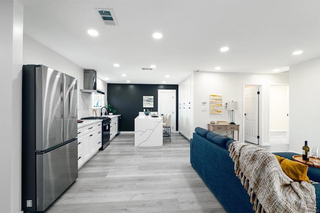 kitchen with gas stove, light hardwood / wood-style flooring, stainless steel refrigerator, wall chimney range hood, and white cabinets