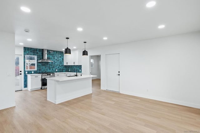 kitchen with pendant lighting, wall chimney exhaust hood, stainless steel stove, white cabinetry, and a center island with sink