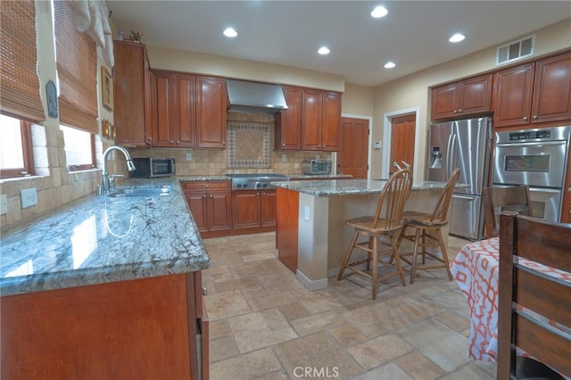 kitchen with wall chimney exhaust hood, sink, a kitchen island, stainless steel appliances, and light stone countertops