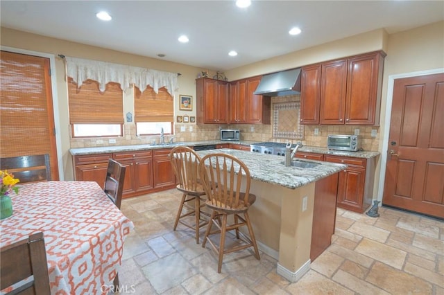 kitchen with backsplash, a kitchen breakfast bar, light stone counters, a center island with sink, and wall chimney exhaust hood