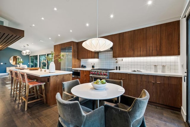 kitchen featuring dark hardwood / wood-style floors, high end stove, decorative backsplash, hanging light fixtures, and a center island with sink