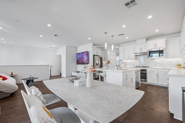 kitchen with dark wood-type flooring, decorative light fixtures, stainless steel appliances, a kitchen island with sink, and white cabinets