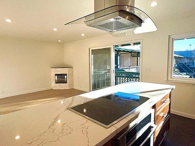 kitchen featuring island range hood, a fireplace, black electric stovetop, stainless steel oven, and light stone countertops