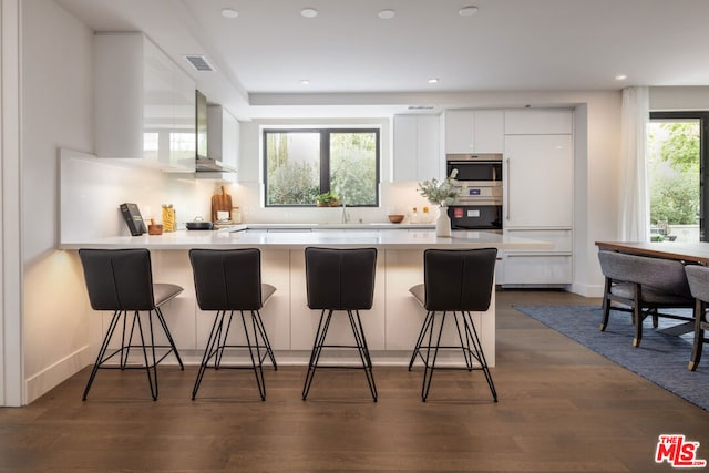 kitchen with dark wood-type flooring, white cabinetry, a kitchen breakfast bar, kitchen peninsula, and wall chimney range hood