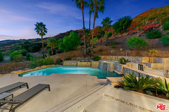 pool at dusk featuring a mountain view, a patio area, and an in ground hot tub