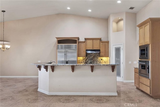 kitchen with light stone counters, built in appliances, decorative light fixtures, a kitchen island with sink, and decorative backsplash