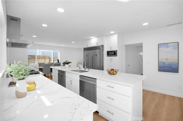 kitchen with a kitchen island with sink, white cabinetry, a sink, light stone countertops, and built in appliances