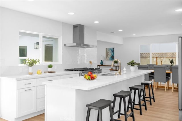 kitchen featuring wall chimney range hood, sink, a kitchen island with sink, backsplash, and white cabinets