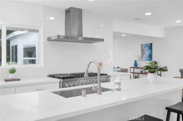 kitchen with white cabinets, light stone countertops, wall chimney range hood, a kitchen bar, and a sink