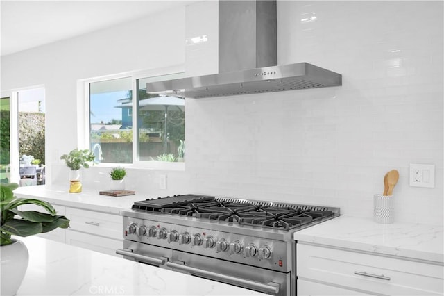 kitchen with wall chimney range hood, tasteful backsplash, light stone counters, white cabinets, and range with two ovens