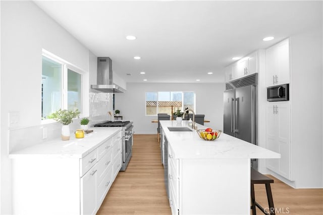 kitchen featuring wall chimney range hood, sink, a kitchen island with sink, built in appliances, and white cabinets