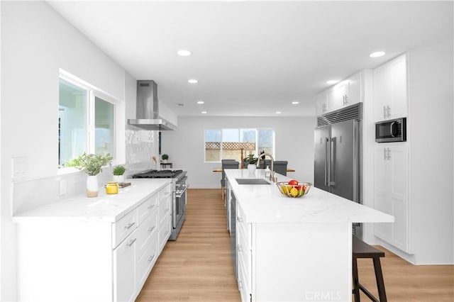 kitchen featuring white cabinetry, a sink, an island with sink, built in appliances, and wall chimney exhaust hood