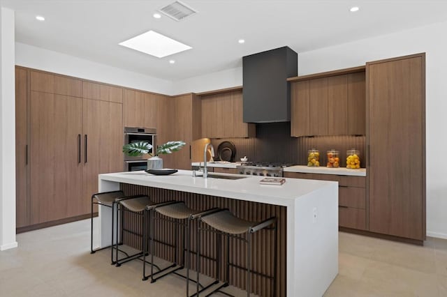 kitchen with sink, wall chimney range hood, range, an island with sink, and decorative backsplash