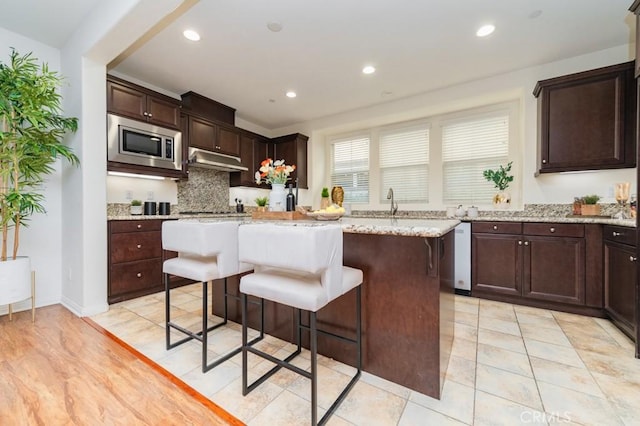 kitchen featuring dark brown cabinets, stainless steel microwave, light stone counters, tasteful backsplash, and a kitchen island