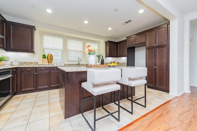 kitchen featuring a kitchen breakfast bar, a center island, stainless steel oven, light stone counters, and dark brown cabinets