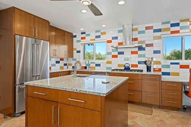 kitchen featuring appliances with stainless steel finishes, backsplash, light stone counters, a center island with sink, and wall chimney range hood