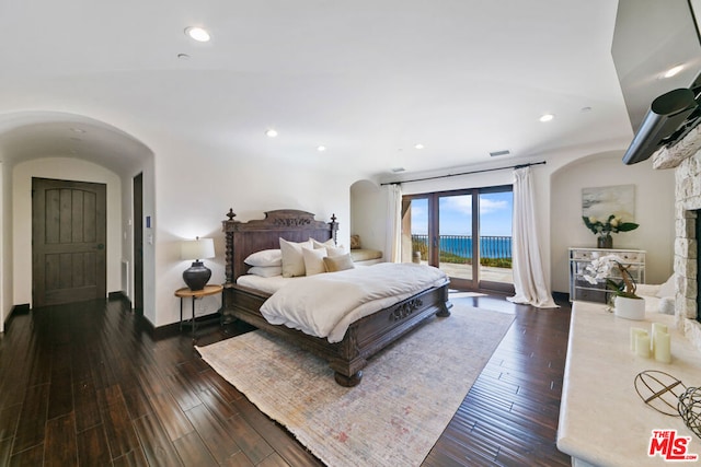 bedroom with dark wood-type flooring, a water view, a stone fireplace, and access to outside