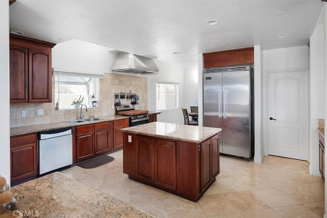 kitchen with appliances with stainless steel finishes, light stone counters, a kitchen island, decorative backsplash, and wall chimney exhaust hood