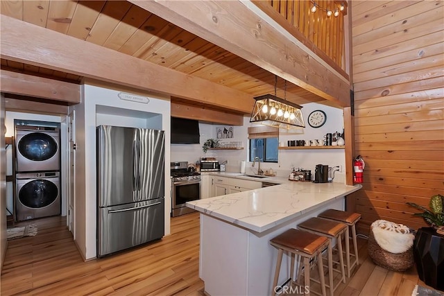 kitchen featuring stacked washing maching and dryer, pendant lighting, wood walls, kitchen peninsula, and stainless steel appliances