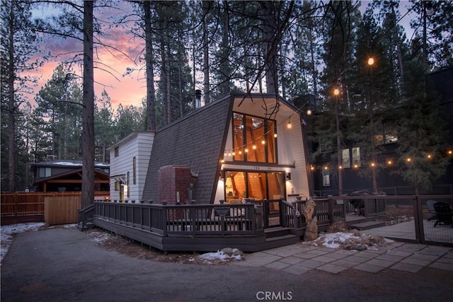 back of property at dusk with roof with shingles, a wooden deck, a gambrel roof, and fence