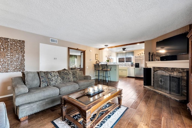 living room featuring ceiling fan, a stone fireplace, dark wood-type flooring, and a textured ceiling