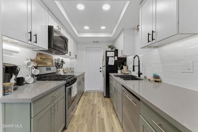 kitchen with a raised ceiling, sink, backsplash, stainless steel appliances, and light wood-type flooring