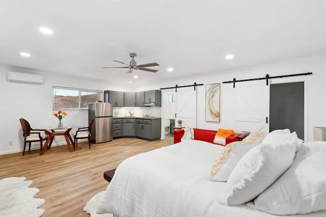 bedroom with a wall mounted air conditioner, stainless steel refrigerator, ceiling fan, a barn door, and light wood-type flooring