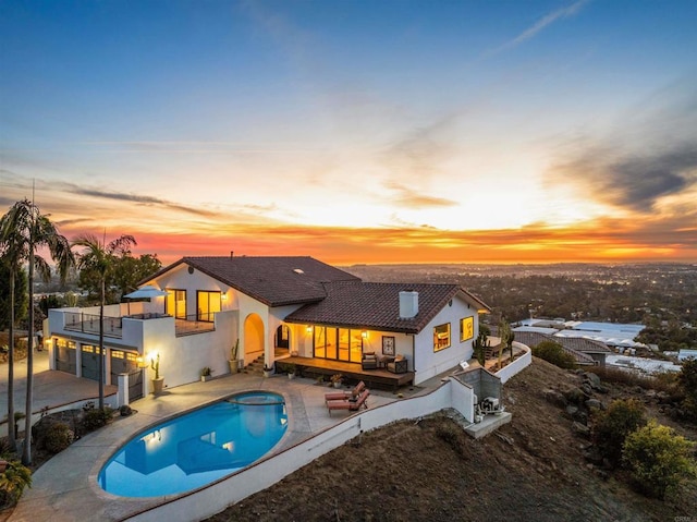 back house at dusk featuring a swimming pool with hot tub, a patio, and a balcony