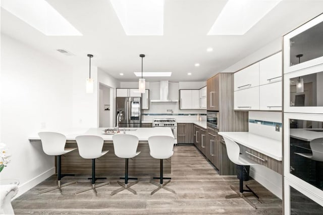 kitchen featuring wall chimney exhaust hood, white cabinetry, hanging light fixtures, a center island with sink, and appliances with stainless steel finishes