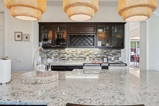 bar with light stone counters, backsplash, pendant lighting, and dark brown cabinets