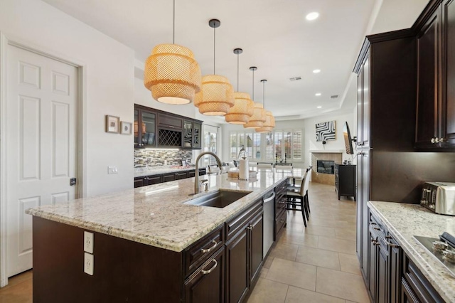 kitchen with sink, a kitchen island with sink, dark brown cabinets, light stone counters, and decorative light fixtures