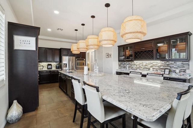 kitchen featuring decorative backsplash, a large island with sink, hanging light fixtures, stainless steel appliances, and dark brown cabinets