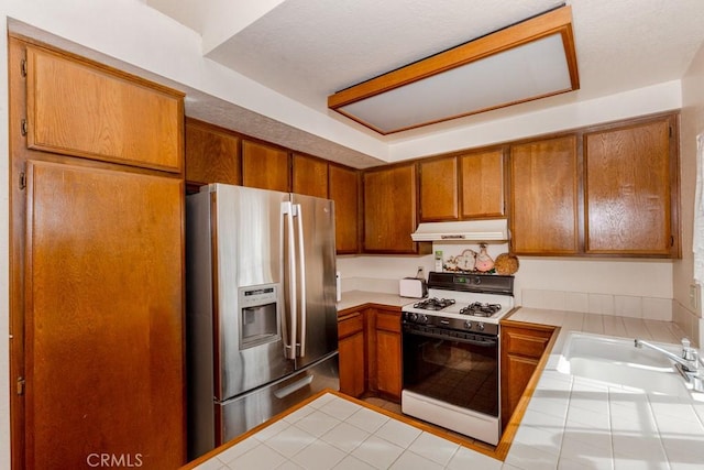 kitchen featuring brown cabinets, under cabinet range hood, stainless steel fridge with ice dispenser, and gas range oven