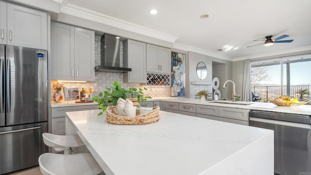 kitchen with wall chimney range hood, crown molding, gray cabinets, appliances with stainless steel finishes, and a kitchen breakfast bar