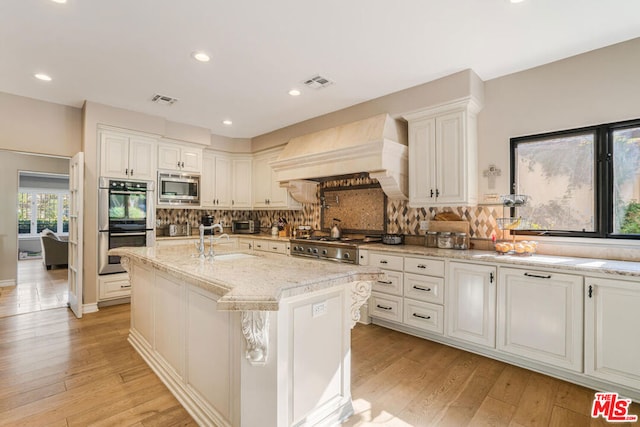 kitchen featuring sink, custom exhaust hood, an island with sink, stainless steel appliances, and light stone countertops