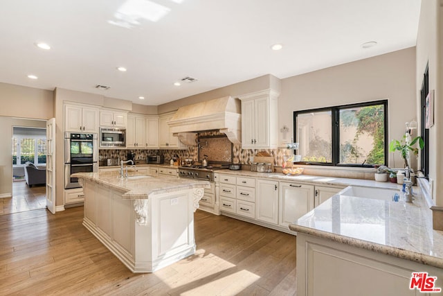 kitchen featuring an island with sink, sink, custom exhaust hood, stainless steel appliances, and light stone countertops