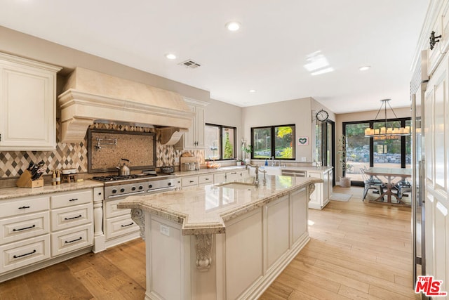 kitchen with premium range hood, stainless steel gas stovetop, sink, a kitchen island with sink, and light stone counters