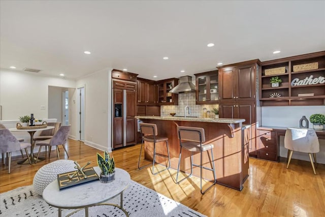 kitchen with a kitchen breakfast bar, a center island, paneled built in fridge, light stone counters, and wall chimney range hood