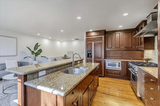 kitchen with sink, a breakfast bar, built in appliances, a center island with sink, and wall chimney exhaust hood