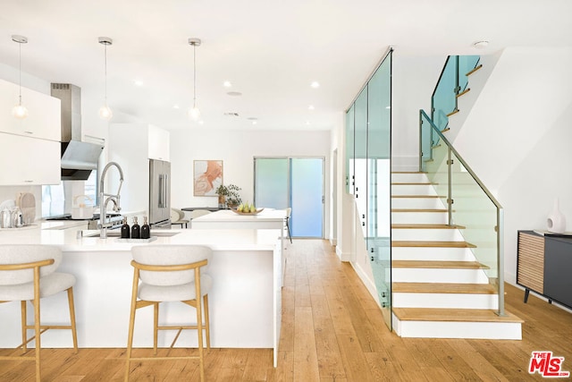 kitchen featuring a breakfast bar area, high end refrigerator, decorative light fixtures, light wood-type flooring, and wall chimney range hood