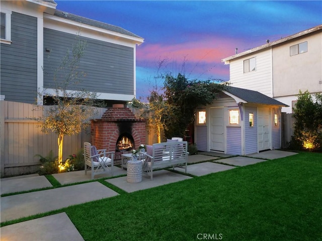 back house at dusk featuring an outdoor brick fireplace, a yard, a patio area, and an outbuilding