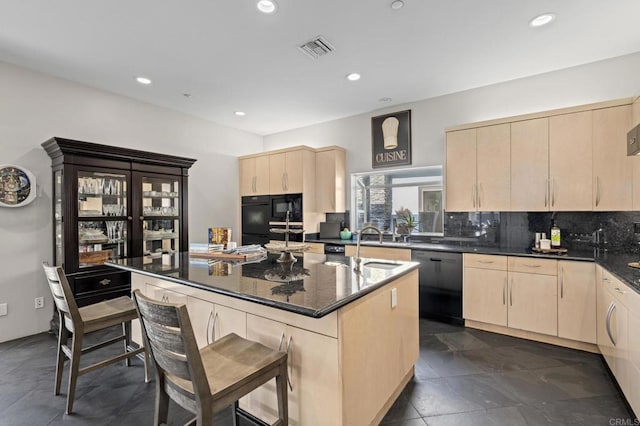 kitchen featuring light brown cabinetry, dishwasher, dark stone countertops, backsplash, and a center island with sink