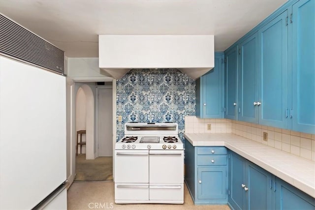 kitchen with blue cabinets, backsplash, tile counters, white appliances, and wall chimney exhaust hood