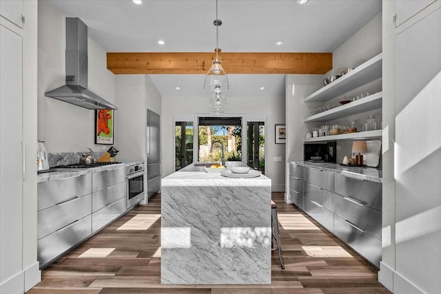 kitchen with wall chimney exhaust hood, beamed ceiling, a center island with sink, appliances with stainless steel finishes, and pendant lighting