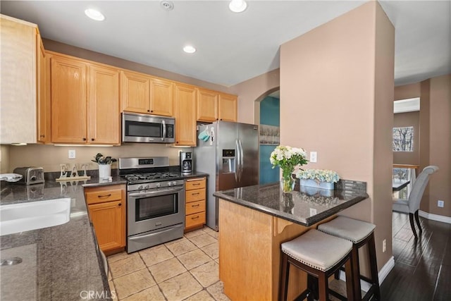 kitchen with a breakfast bar, sink, dark stone counters, stainless steel appliances, and light brown cabinets