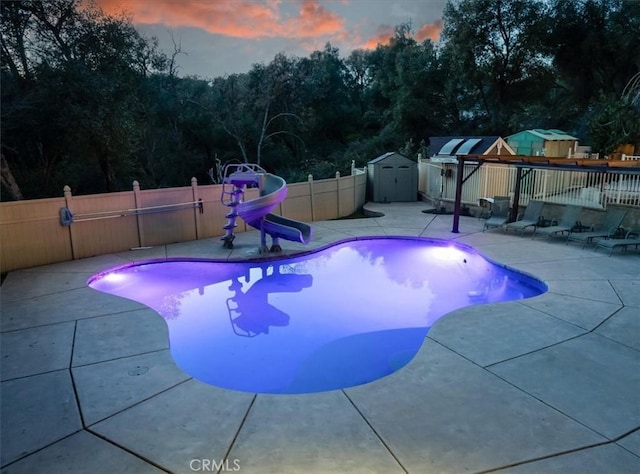 pool at dusk featuring a storage shed, a water slide, and a patio area