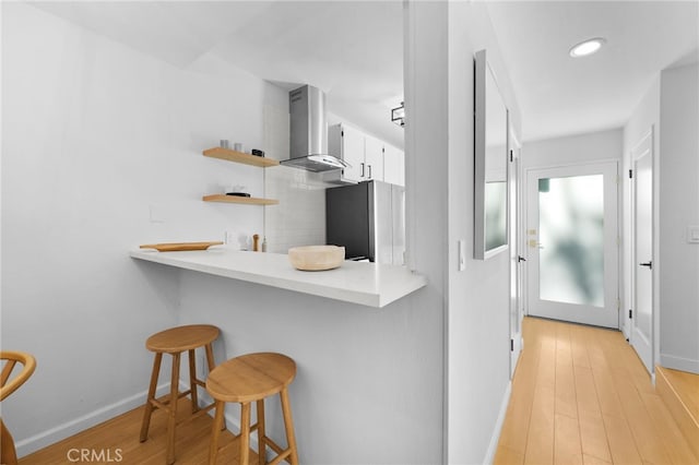 kitchen featuring stainless steel refrigerator, a breakfast bar, white cabinetry, light hardwood / wood-style floors, and wall chimney exhaust hood