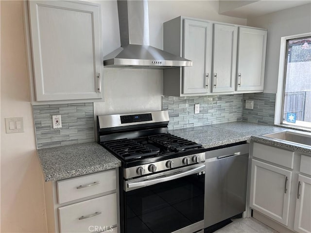 kitchen featuring appliances with stainless steel finishes, white cabinetry, backsplash, light stone countertops, and wall chimney range hood